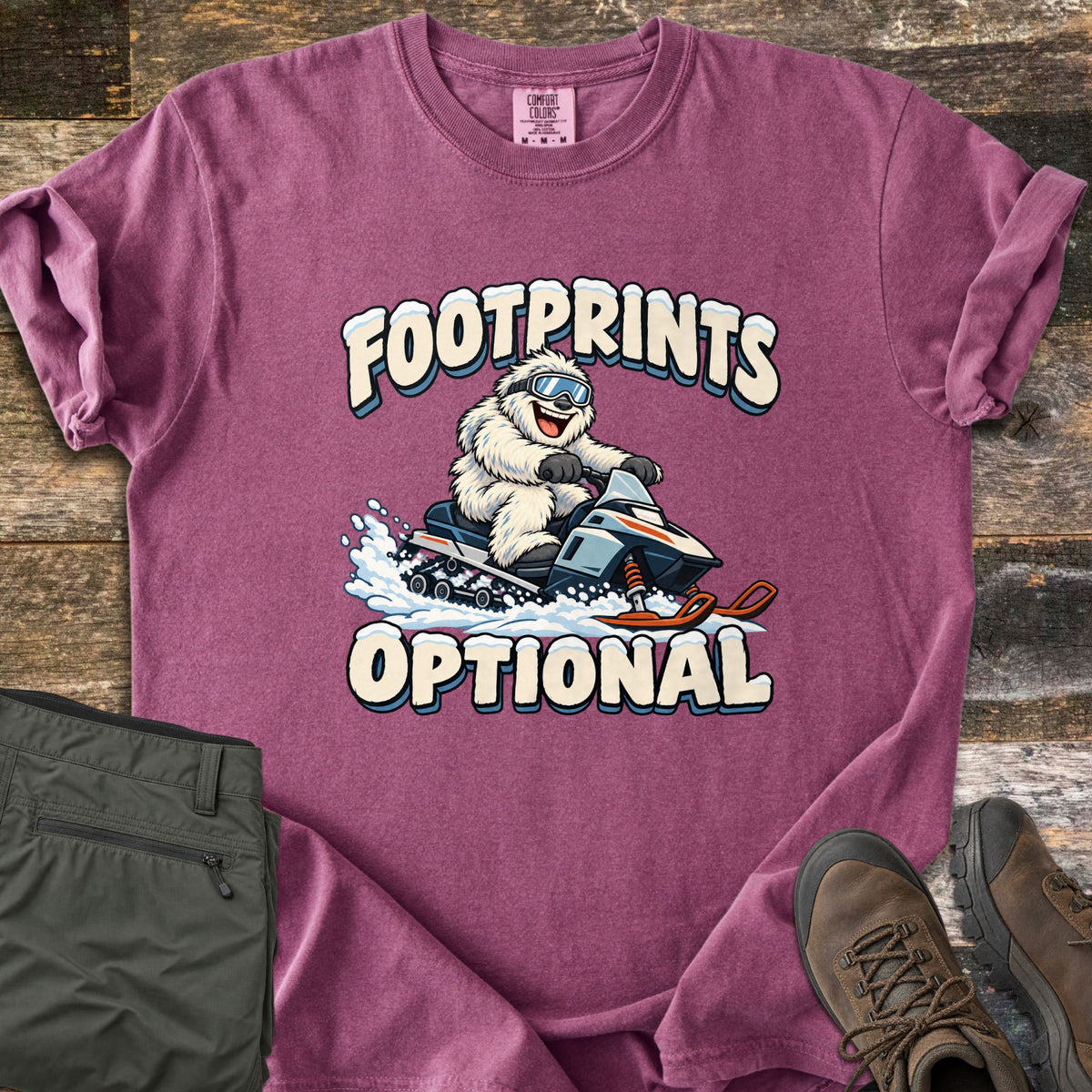 Footprints Optional Comfort Colors T-shirt in Berry, laid flat. A grinning white yeti in ski goggles rides a snowmobile through spraying snow between the snow-capped words Footprints Optional. The sleeves are cuffed, on weathered wood with folded olive pants and brown hiking boots.