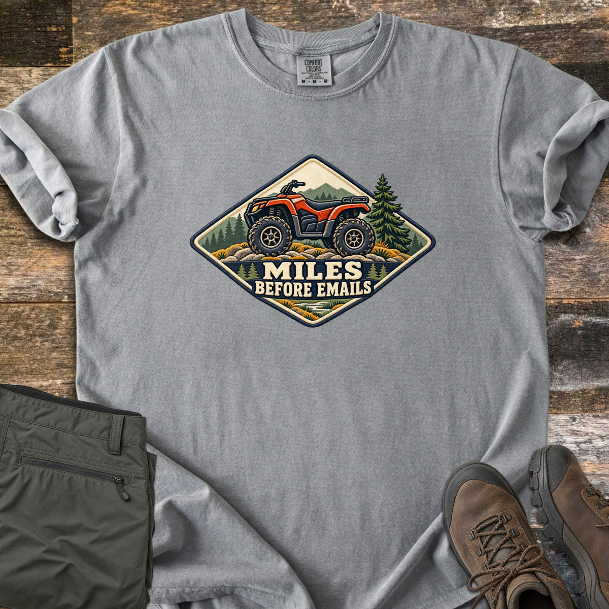 Miles Before Emails Comfort Colors T-shirt in Grey, laid flat. A diamond-shaped patch shows a red ATV on rocks with pine trees and mountains above the words Miles Before Emails. Sleeves rolled, on weathered wood with green pants and brown hiking boots.