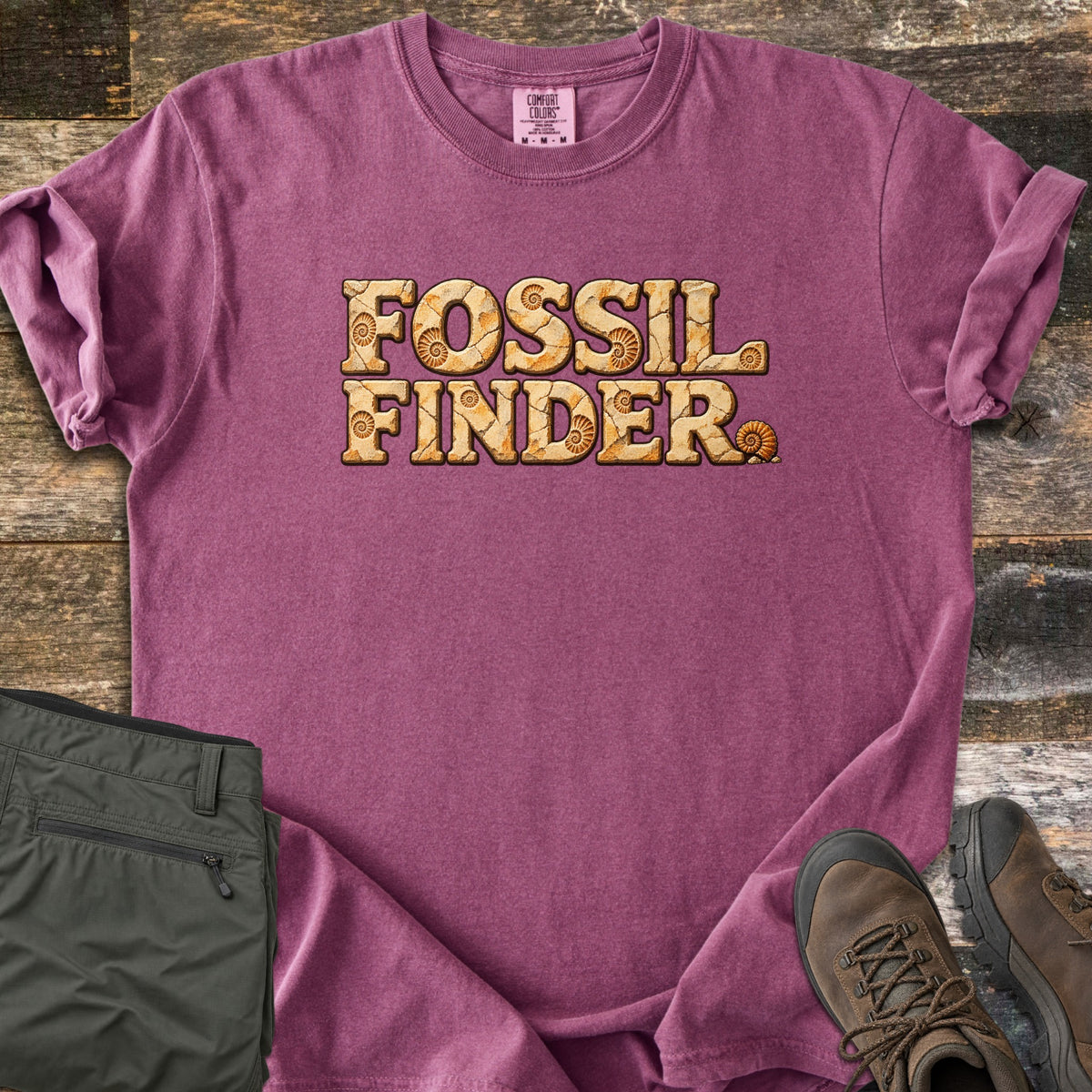 Fossil Finder Comfort Colors T-shirt in Berry, laid flat. The words Fossil Finder are printed in cracked tan stone letters with ammonite shell fossils, plus a small ammonite after the last letter. Sleeves are rolled, on a rustic wood floor with olive cargo pants and brown hiking boots.
