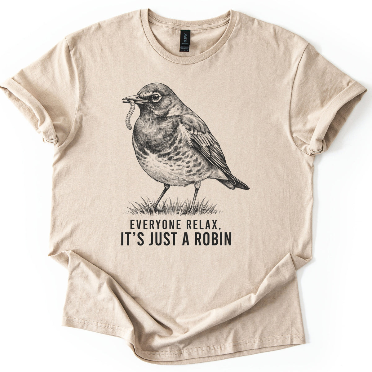 Everyone Relax It's Just A Robin Comfort Colors T-shirt in Sand, laid flat. A black sketch-style drawing of a robin holding a worm in its beak, standing on a tuft of grass, above the words Everyone Relax, It's Just A Robin. The sleeves are rolled on a white background.