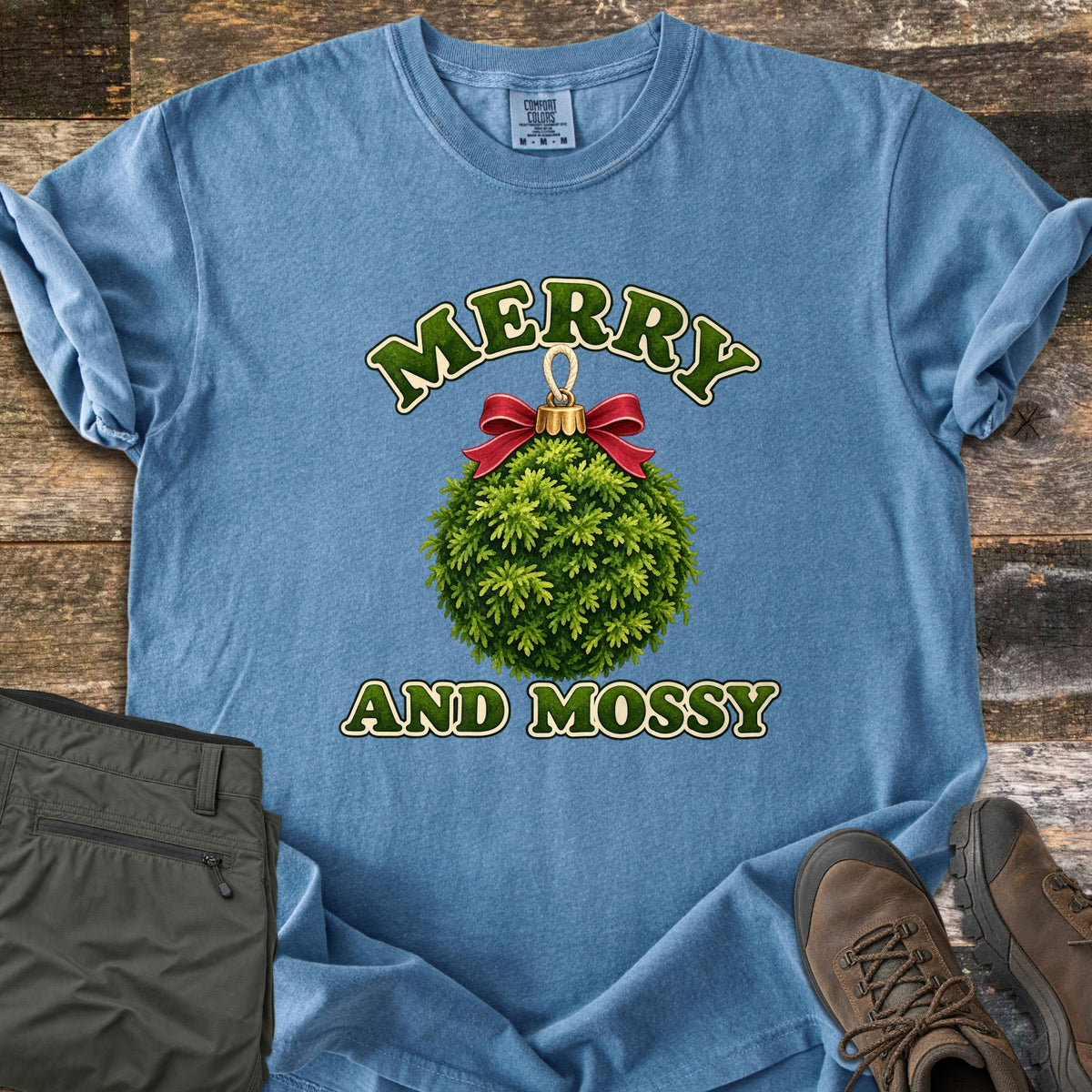 Merry and Mossy Comfort Colors T-shirt in Blue Jean, laid flat. A round Christmas ornament made of green moss, topped with a gold cap and red bow, sits between the words Merry and Mossy in green lettering. Olive hiking pants and brown hiking boots lie beside it on weathered wood.