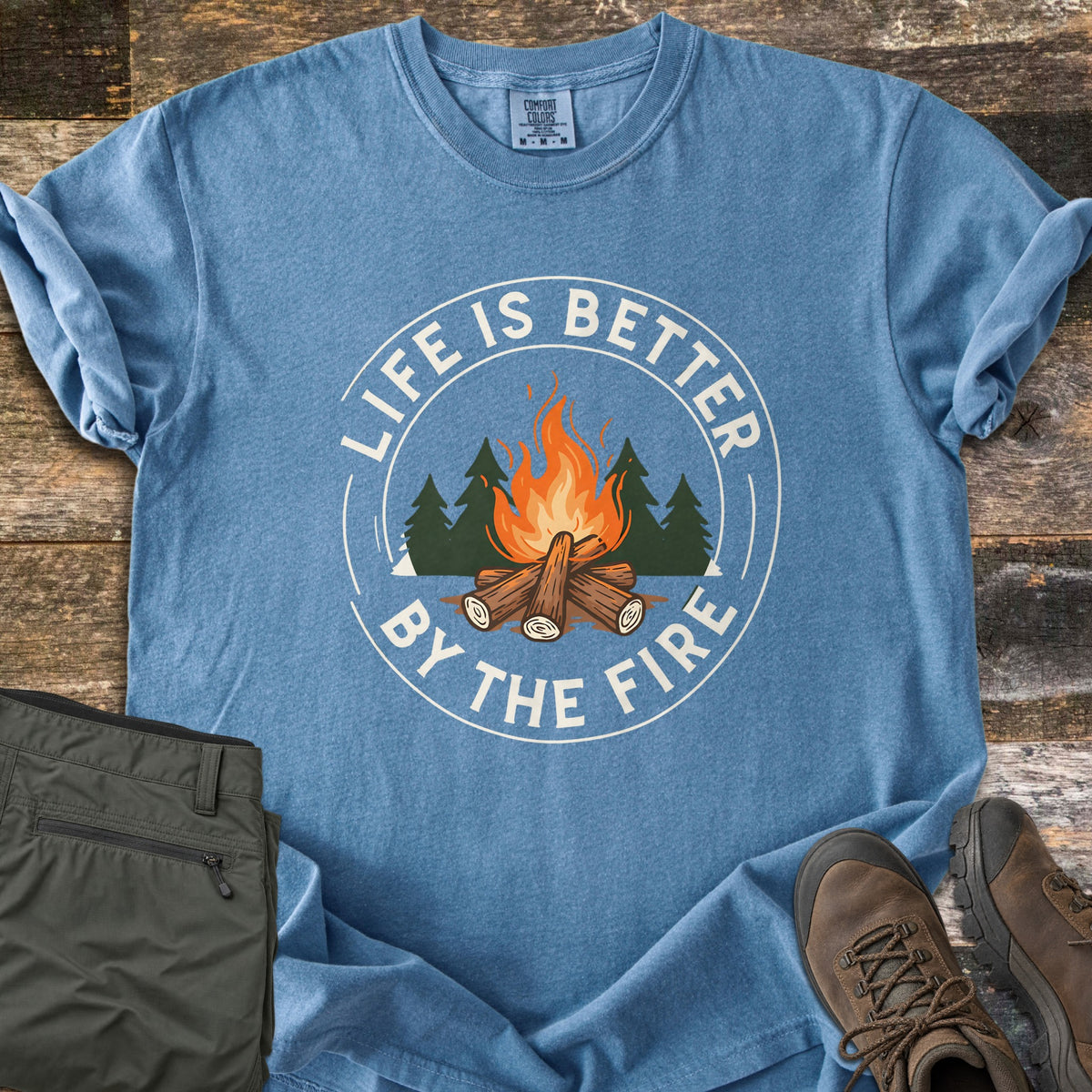 Life Is Better By The Fire Ring Comfort Colors T-shirt in Blue Jean, laid flat. A round badge shows an orange log campfire in front of dark green pine trees, ringed in white by the words Life Is Better By The Fire. Olive hiking pants and brown hiking boots lie beside it on weathered wood planks.