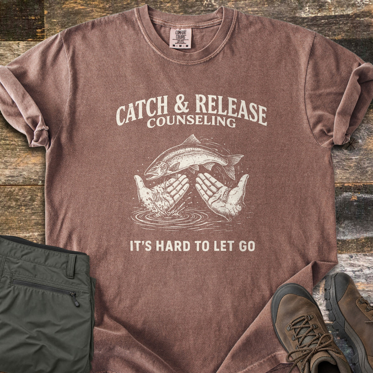 Catch & Release Counseling Comfort Colors T-shirt in Espresso, laid flat. A trout leaps between two open hands over rippling water under the words Catch & Release Counseling, with It's Hard to Let Go below, in cream ink. Folded olive hiking pants and brown boots sit on weathered wood.