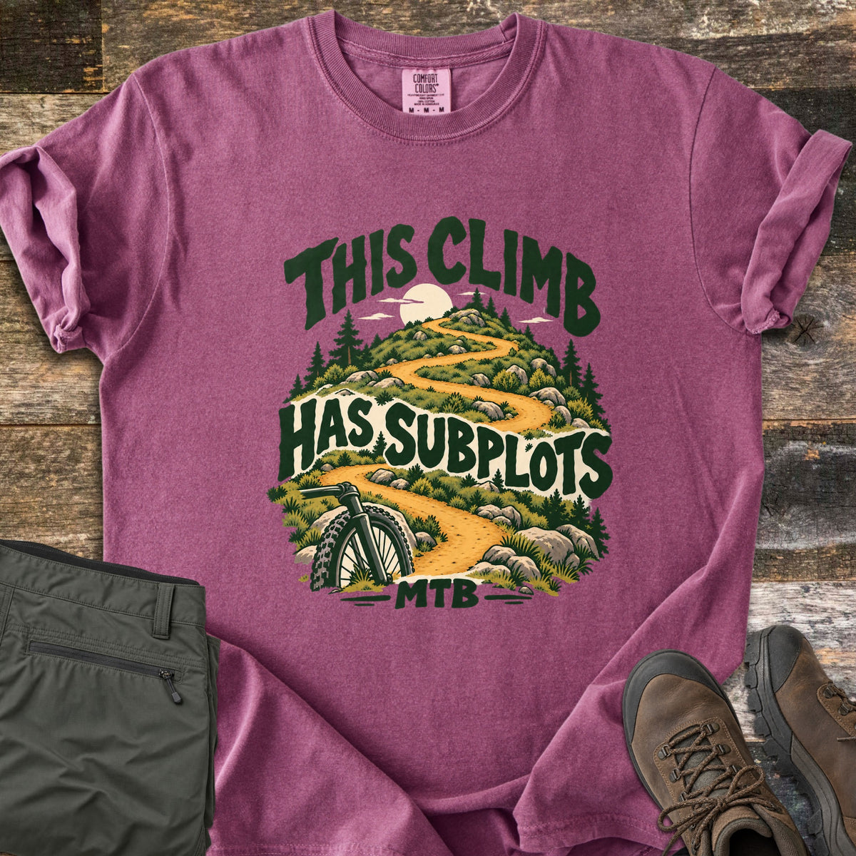 This Climb Has Subplots Comfort Colors T-shirt in Berry, laid flat. Dark green words This Climb Has Subplots MTB surround a winding dirt trail up a rocky hill with pines, a pale sun, and a mountain bike wheel. Folded olive pants and brown hiking boots lie on rustic wood.