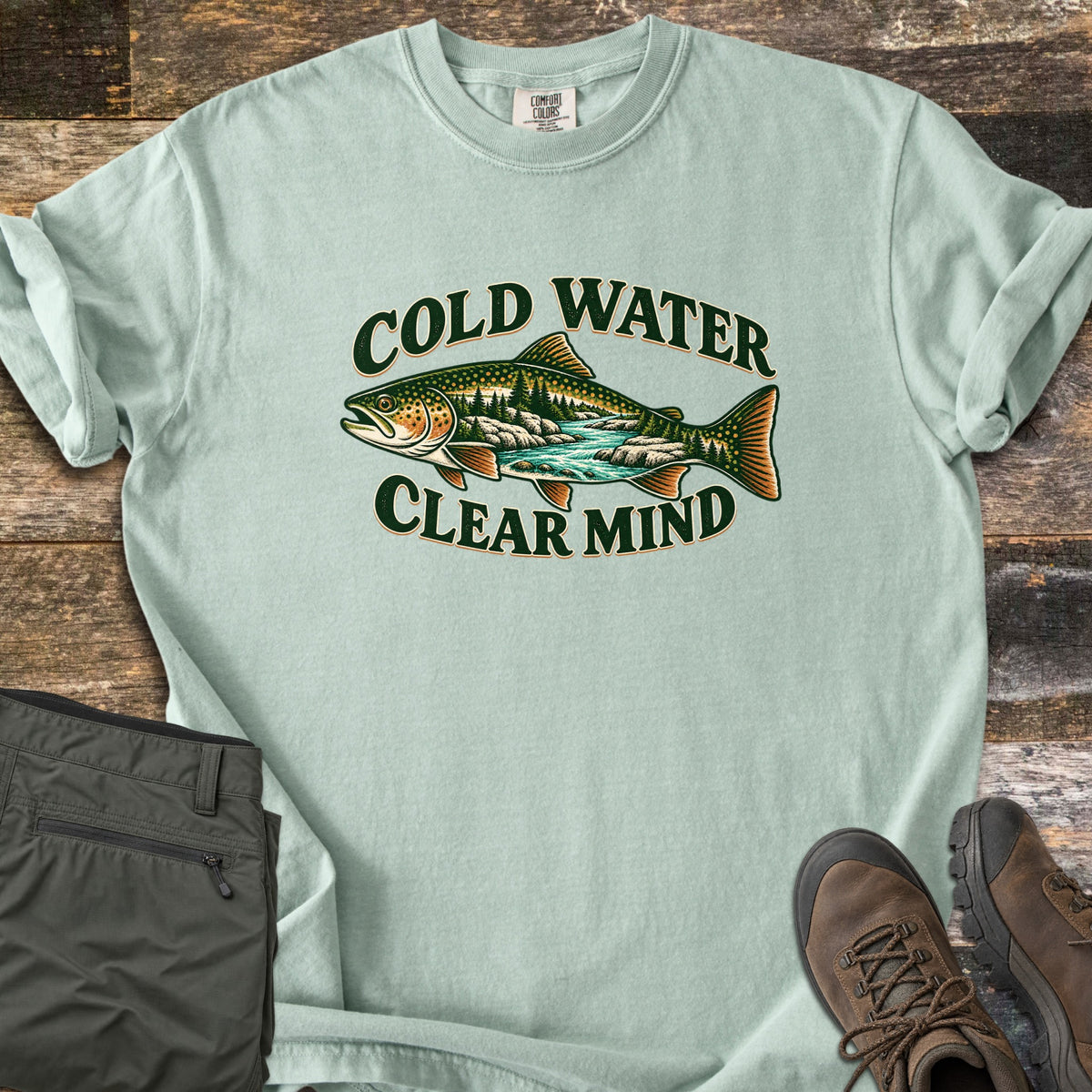 Cold Water Clear Mind Comfort Colors T-shirt in Bay, laid flat. A spotted trout filled with pine trees, rocks and a turquoise river is framed by the dark green words Cold Water above and Clear Mind below. Olive hiking pants and brown boots sit beside it on rustic wood planks.
