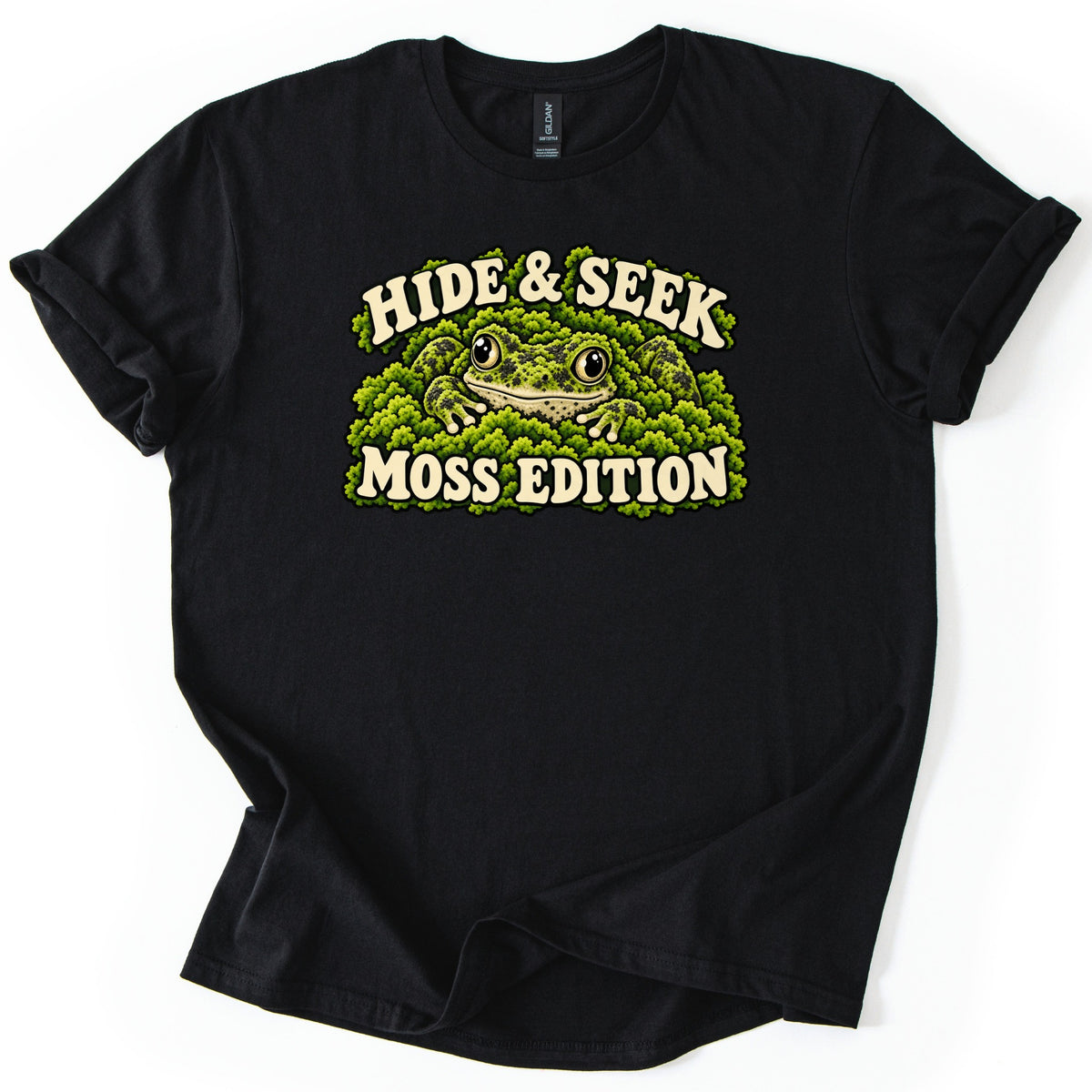 Hide & Seek Moss Edition Comfort Colors T-shirt in Black, laid flat. A spotted green frog with big round eyes hides in thick green moss, framed by the cream words Hide & Seek above and Moss Edition below. The shirt lies on a white surface with its sleeves rolled.