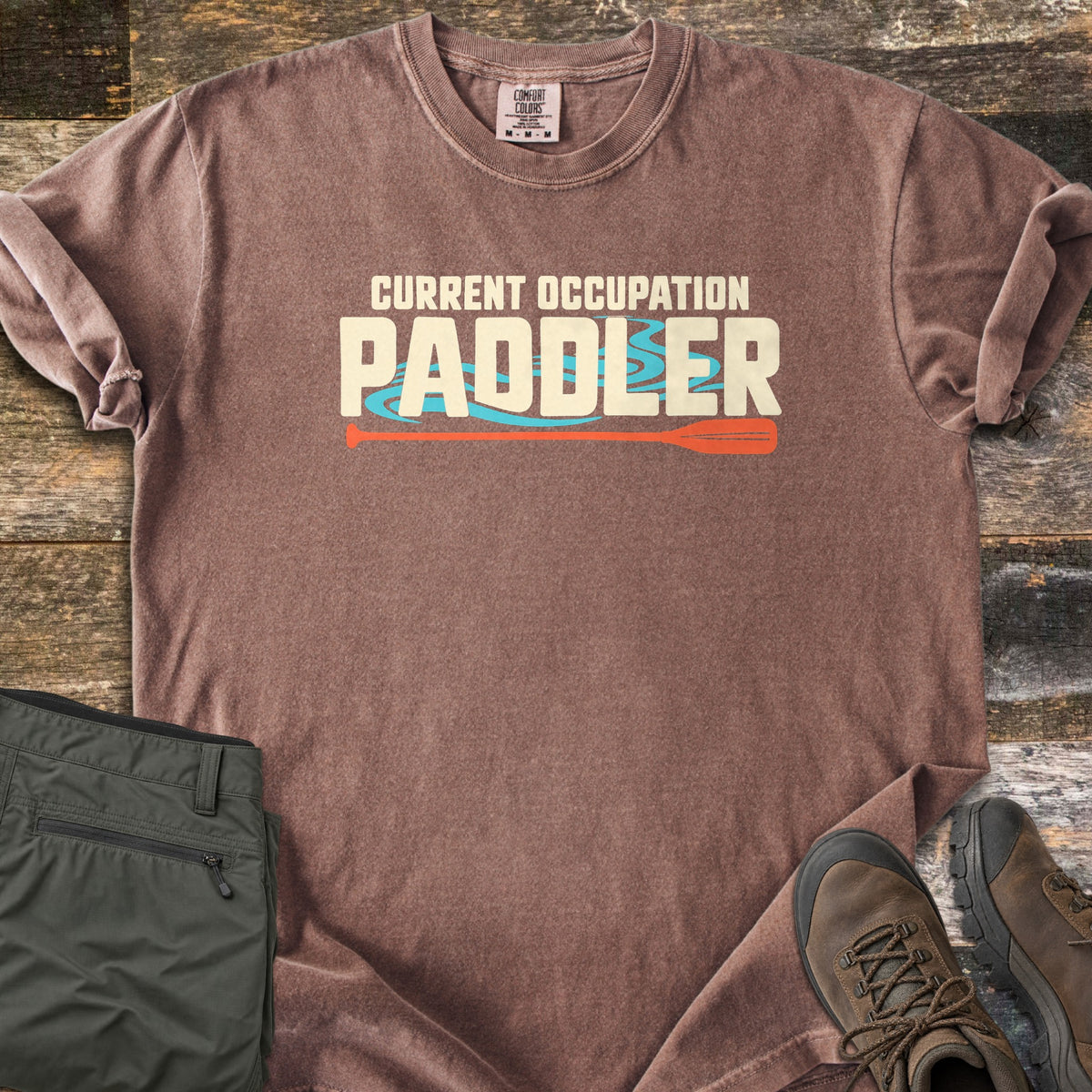 Current Occupation Paddler Comfort Colors T-shirt in Espresso, laid flat. Cream block letters read Current Occupation Paddler over turquoise water swirls, with an orange canoe paddle beneath. Sleeves are rolled on a weathered wood surface beside olive hiking pants and brown hiking boots.