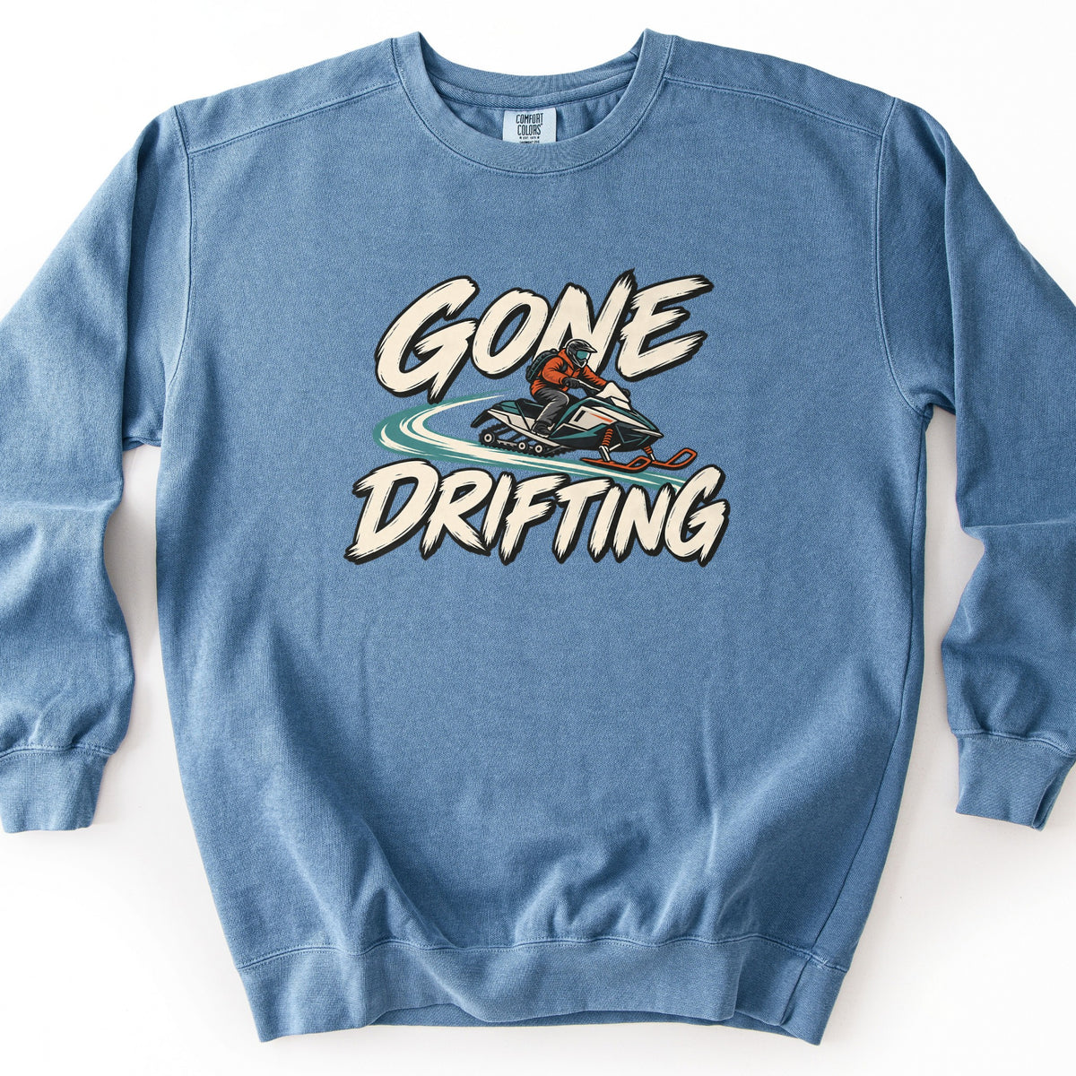 Gone Drifting Comfort Colors crewneck sweatshirt in Blue Jean, laid flat. A rider in an orange jacket and helmet drifts a teal and white snowmobile, kicking up a teal snow trail, between the words Gone Drifting in cream brush lettering. Shown on a white background.