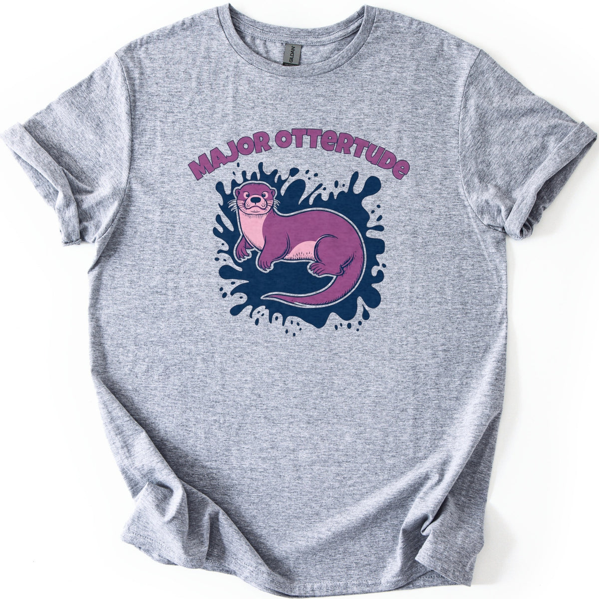 Major Ottertude Comfort Colors T-shirt in Sport Grey, laid flat. Arched purple letters reading MAJOR OTTERTUDE sit above a purple cartoon otter with a pink chest lying on a navy blue paint splash. The shirt has cuffed sleeves and lies on a white background.
