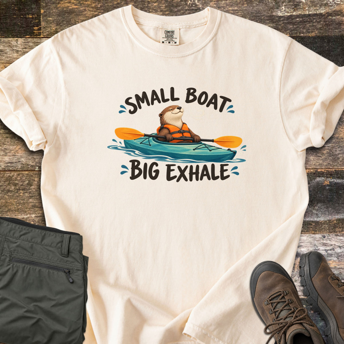 Small Boat Big Exhale Comfort Colors T-shirt in Ivory, laid flat. A smiling cartoon otter in an orange life vest paddles a teal kayak between the words Small Boat Big Exhale. The shirt lies on weathered wood beside folded olive hiking pants and brown hiking boots.