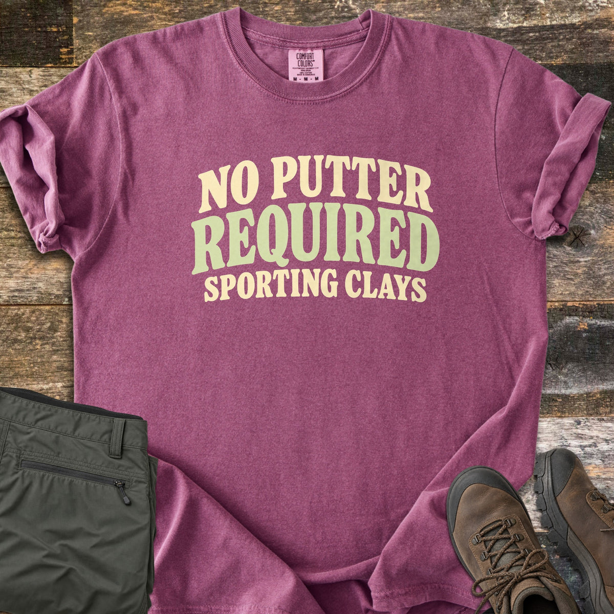 No Putter Required Comfort Colors T-shirt in Berry, laid flat. Wavy block letters read No Putter Required in cream and sage green, with Sporting Clays below. Sleeves are rolled on a rustic wood surface beside folded olive cargo pants and brown leather hiking boots.