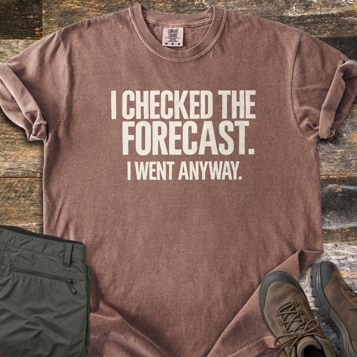 Checked The Forecast Comfort Colors T-shirt in Espresso, laid flat. Off-white block letters read I CHECKED THE FORECAST. I WENT ANYWAY. The sleeves are cuffed, with folded olive hiking pants and brown hiking boots beside it on weathered wood planks.