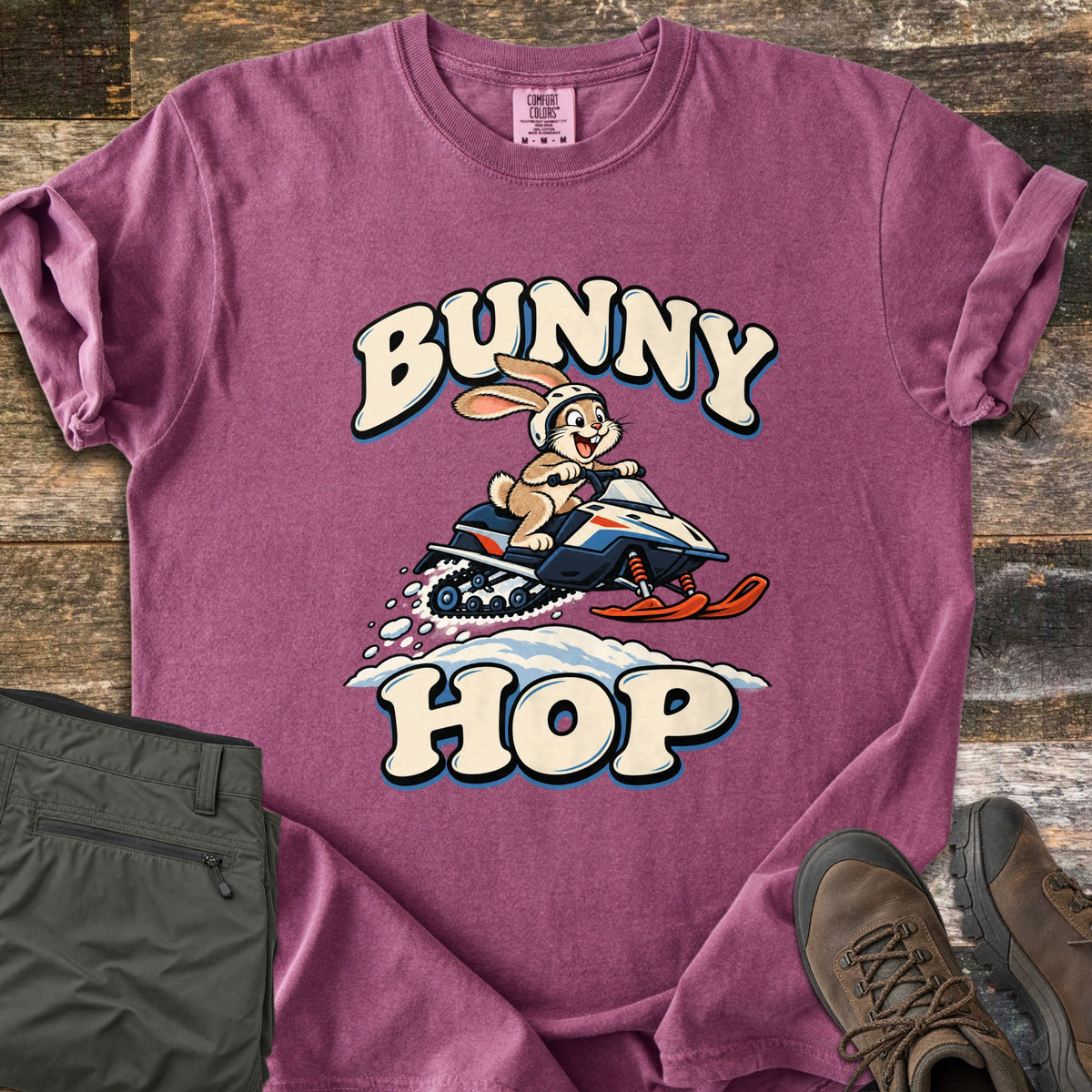 Bunny Hop Comfort Colors T-shirt in Berry, laid flat. A cartoon bunny in a white helmet rides a navy, white and orange snowmobile over a snowbank between the words Bunny Hop. Folded green pants and brown hiking boots lie beside it on a wood floor.