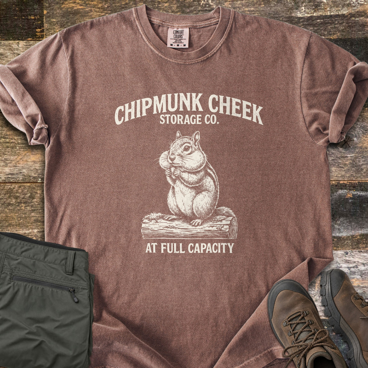 Chipmunk Cheek Storage Co Comfort Colors T-shirt in Espresso, laid flat. Cream print of a chipmunk with full cheeks sitting on a log, under the words Chipmunk Cheek Storage Co. and above At Full Capacity. Rolled sleeves, on weathered wood with olive pants and brown hiking boots.