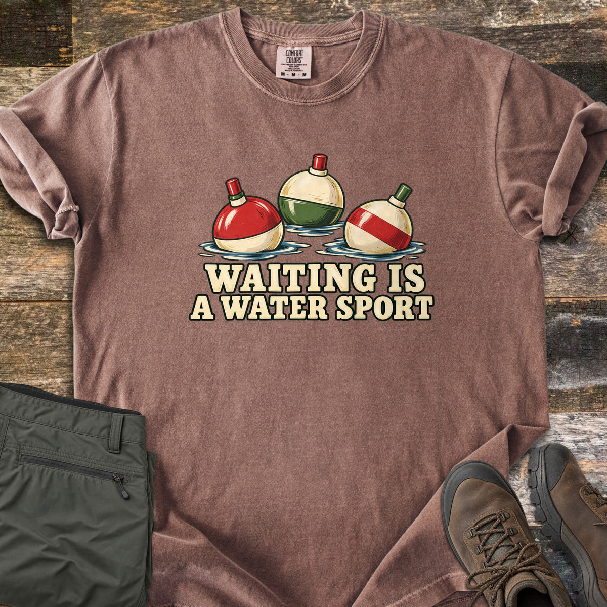 Waiting Is a Water Sport Comfort Colors T-shirt in Espresso, laid flat. Three red, green and cream fishing bobbers float in water above the words Waiting Is a Water Sport. The shirt lies on weathered wood beside folded olive pants and brown hiking boots.