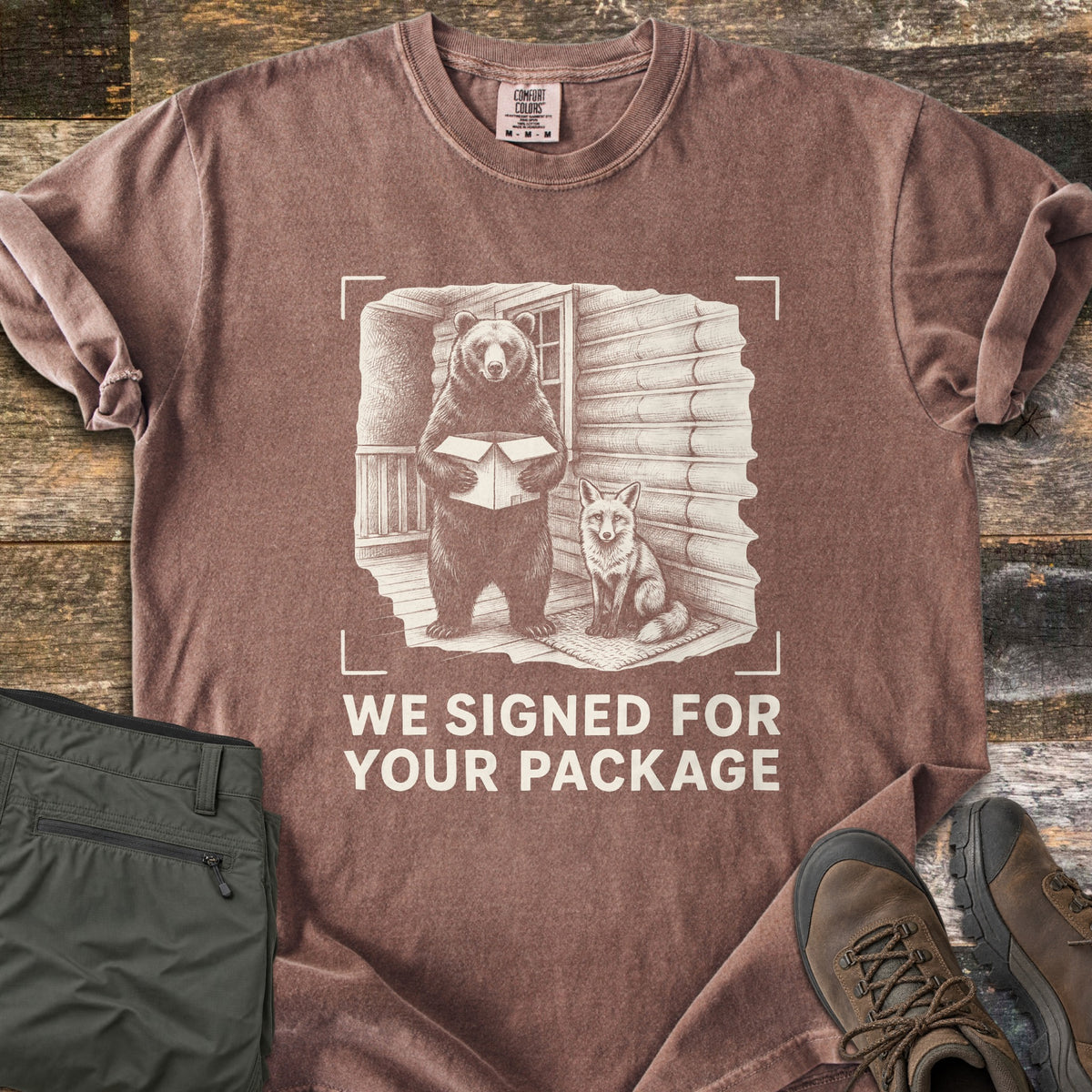 We Signed For Your Package Comfort Colors T-shirt in Espresso, laid flat. A cream-ink sketch shows a bear holding an open box beside a seated fox on a log cabin porch, framed by camera corner marks above the words We Signed For Your Package. Olive pants and brown hiking boots sit on wood planks.