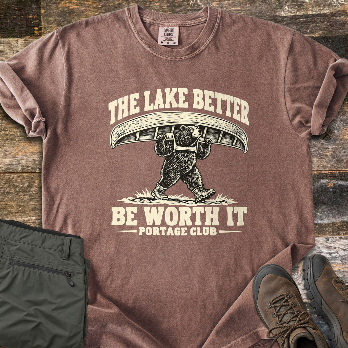 The Lake Better Be Worth It Comfort Colors T-shirt in Espresso, laid flat. A bear in hiking boots carries a canoe overhead between the words The Lake Better Be Worth It, with Portage Club below, printed in cream. Shown on weathered wood with folded green pants and brown hiking boots.