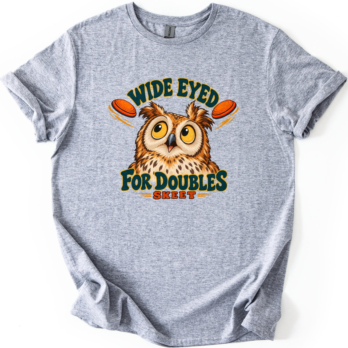 Wide Eyed for Doubles Comfort Colors T-shirt in Sport Grey, laid flat. A wide-eyed cartoon owl looks up between two flying orange clay targets, with Wide Eyed arched above and For Doubles and Skeet below. The shirt is spread on a white surface with rolled sleeves.