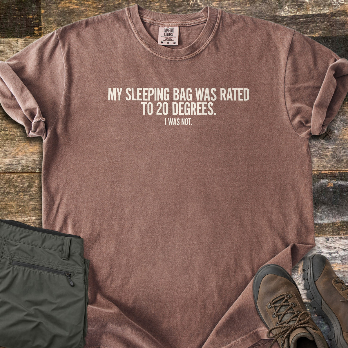 Rated To 20 Degrees Comfort Colors T-shirt in Espresso, laid flat. Cream text across the chest reads My Sleeping Bag Was Rated To 20 Degrees. I Was Not. The shirt has rolled sleeves and lies on weathered wood planks beside folded olive hiking pants and brown hiking boots.