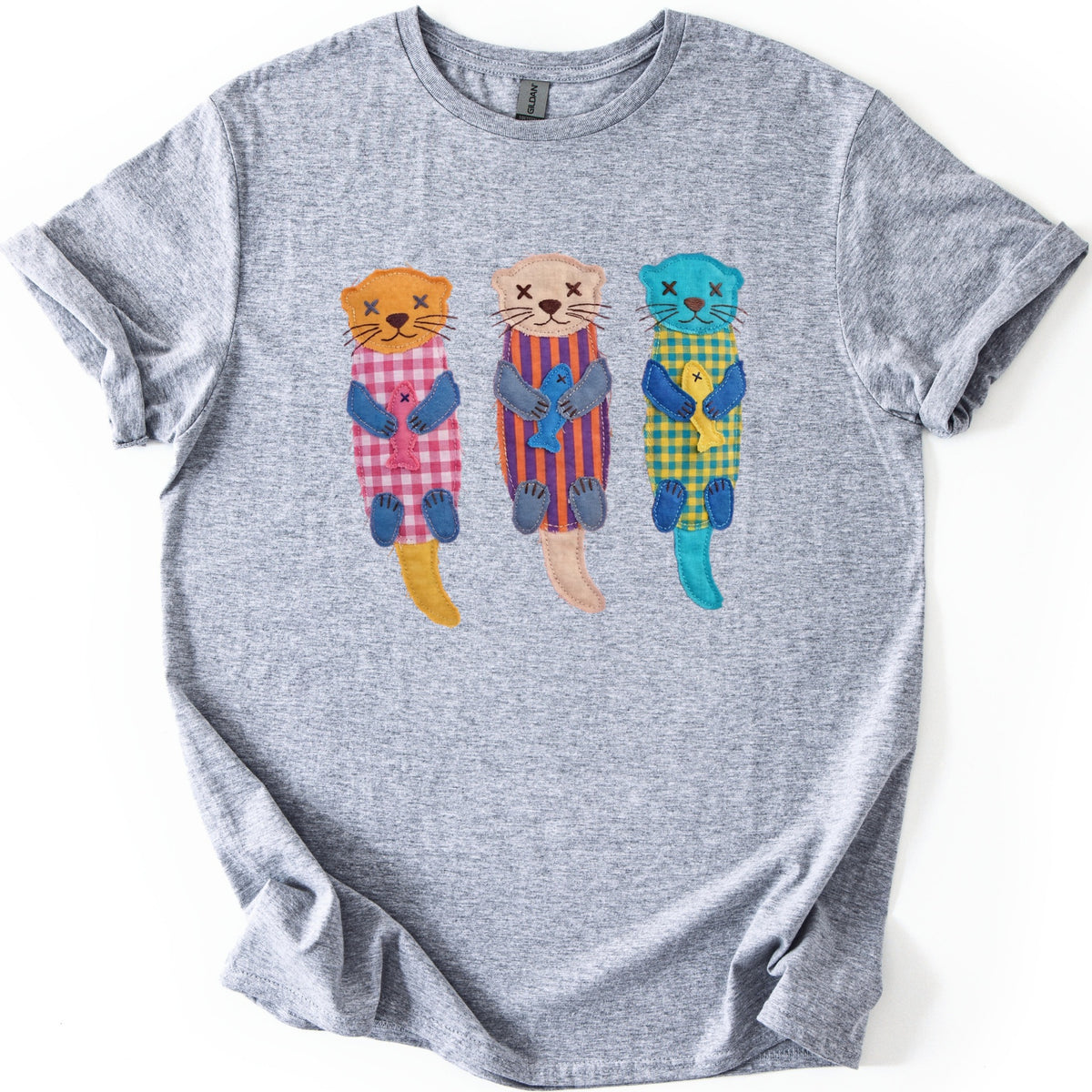 Patchwork Otter Comfort Colors T-shirt in Sport Grey, laid flat. Three appliqué-style otters with X-stitched eyes and blue paws hold fish: orange in pink gingham, beige in purple and orange stripes, turquoise in yellow gingham. No words. Rolled sleeves on a white background.
