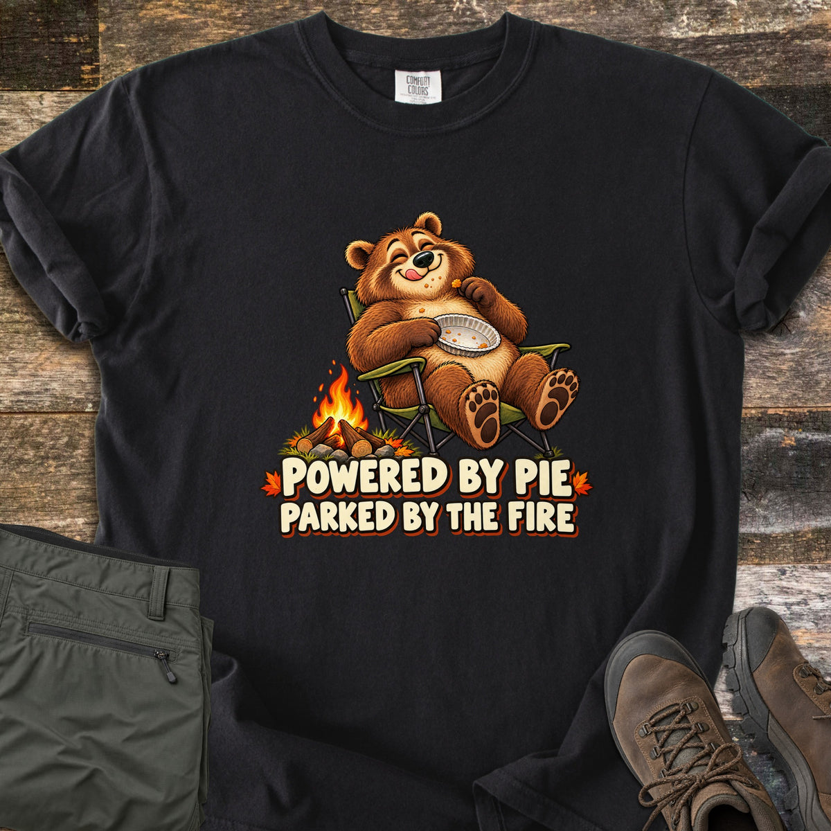 Powered by Pie Parked by the Fire Comfort Colors T-shirt in Black, laid flat. A cartoon brown bear lounges in a green camp chair holding an empty pie tin beside a campfire, above the words Powered by Pie Parked by the Fire. Olive pants and brown hiking boots lie on weathered wood.