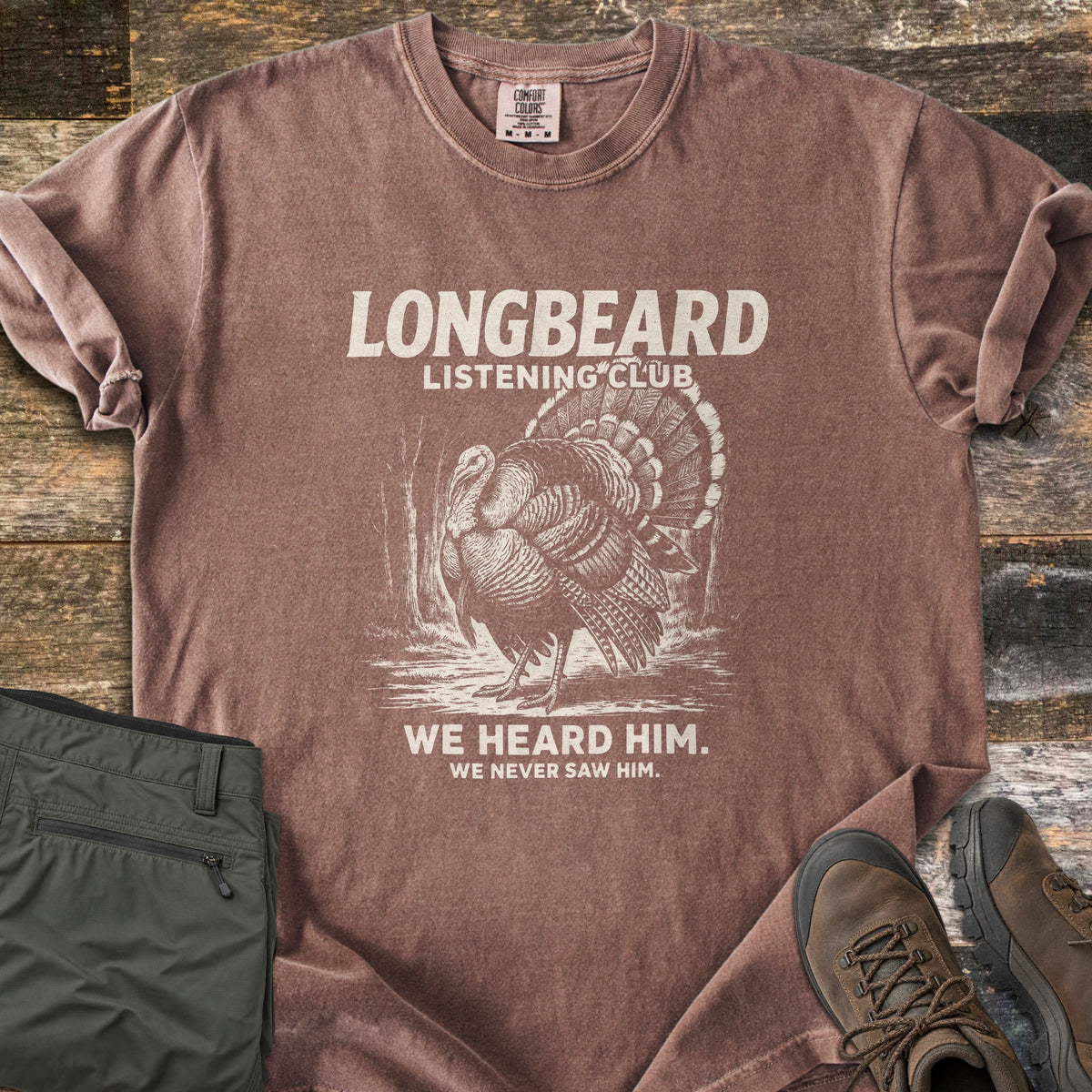 Longbeard Listening Club Comfort Colors T-shirt in Espresso, laid flat. Longbeard Listening Club is printed in cream above a sketched wild turkey fanning its tail among trees, with We heard him. We never saw him. below. Olive hiking pants and brown boots sit on weathered wood.