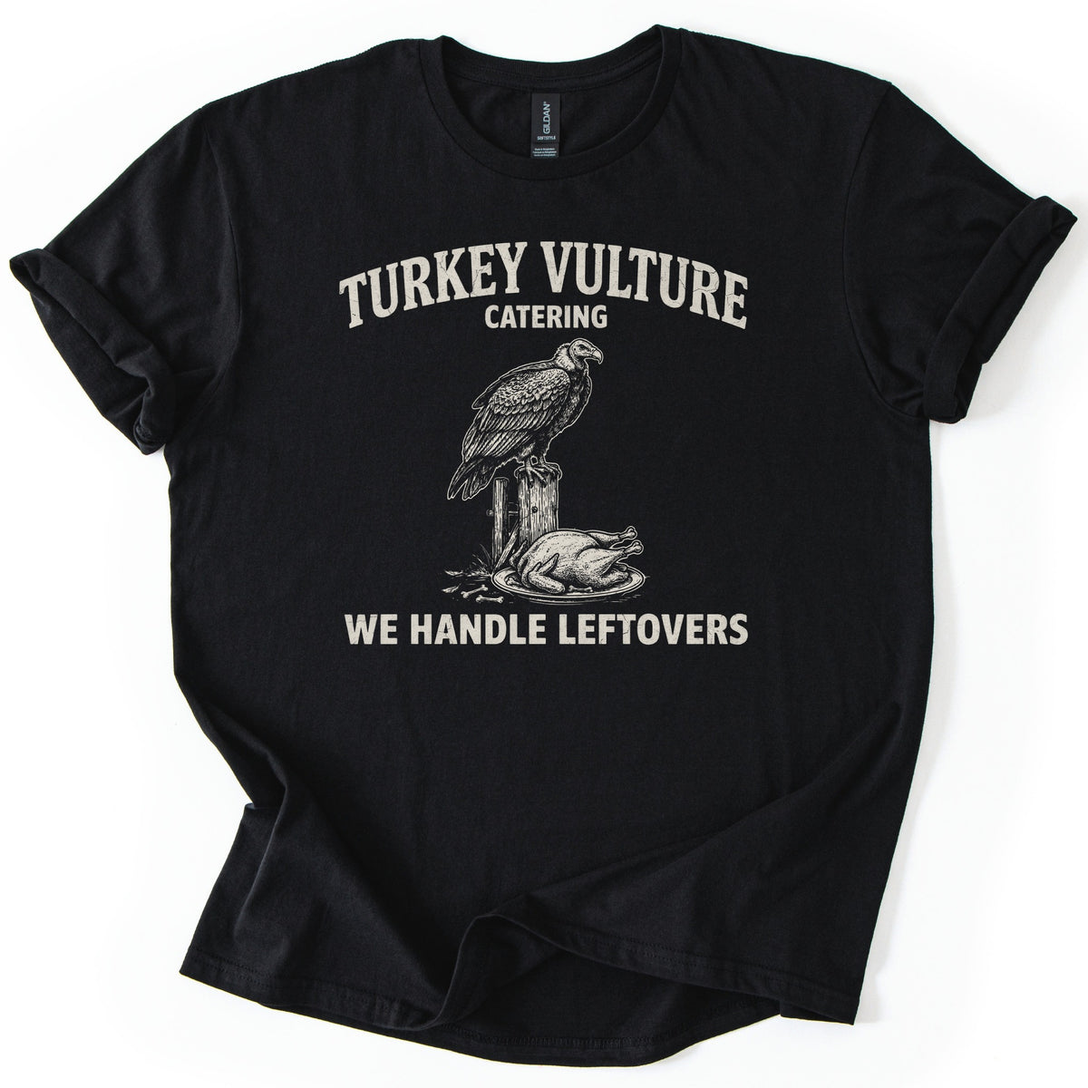 Turkey Vulture Catering Comfort Colors T-shirt in Black, laid flat. A sketched vulture perched on a wooden fence post beside a roast turkey on a platter, under the words Turkey Vulture Catering and above We Handle Leftovers, in cream ink. The sleeves are rolled on a white background.