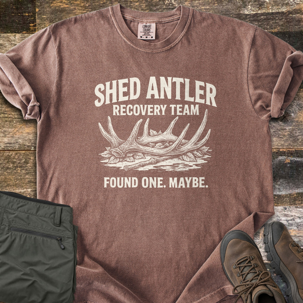 Shed Antler Recovery Team Comfort Colors T-shirt in Espresso, laid flat. A sketched shed antler rests on leaves beside a stick under the words Shed Antler Recovery Team, with Found One. Maybe. below. Olive pants and brown hiking boots sit beside it on weathered wood.