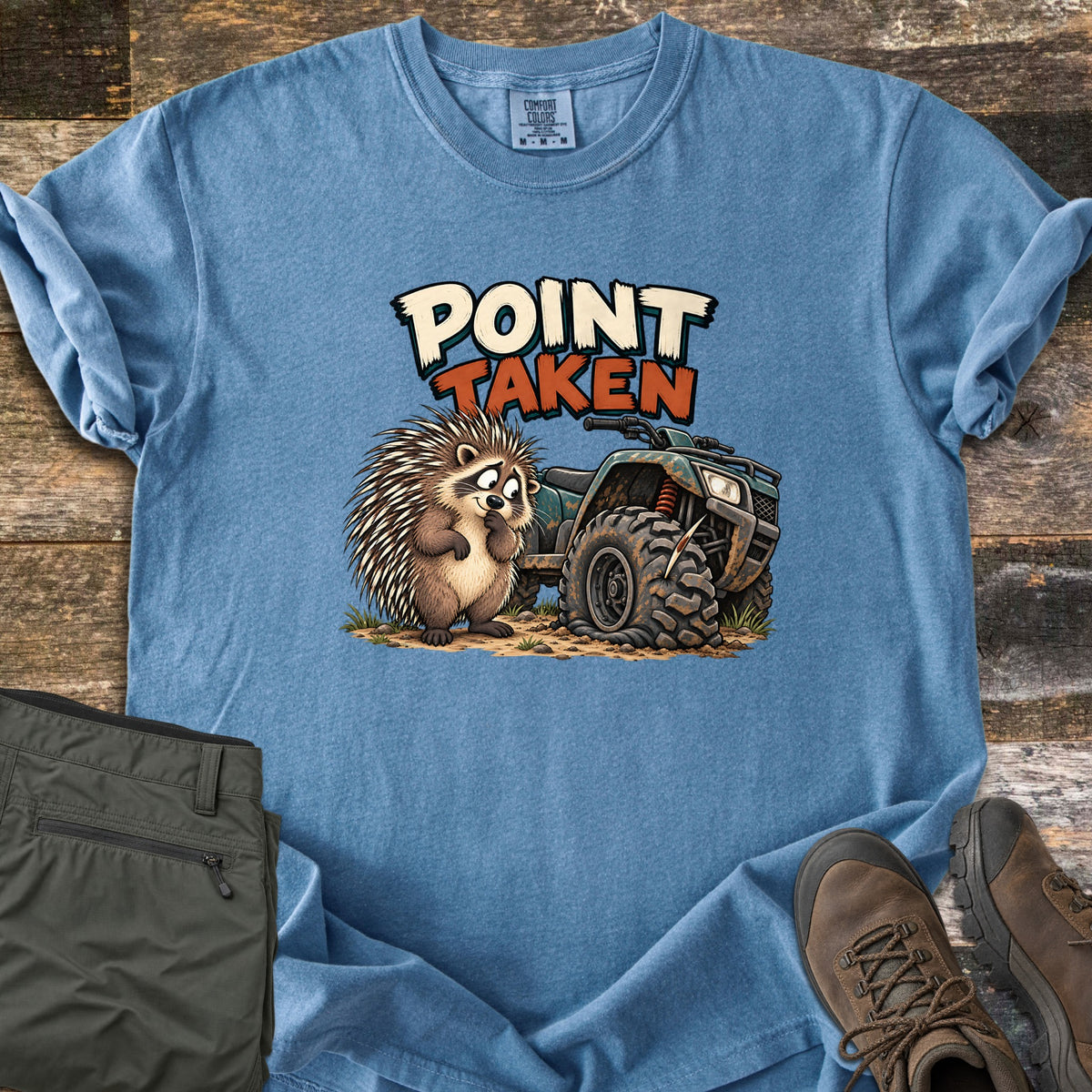 Point Taken Comfort Colors T-shirt in Blue Jean, laid flat. A cartoon porcupine looks sheepishly at a muddy ATV with a flat front tire pierced by a quill, under the words Point Taken. Folded olive pants and brown hiking boots sit beside it on weathered wood.