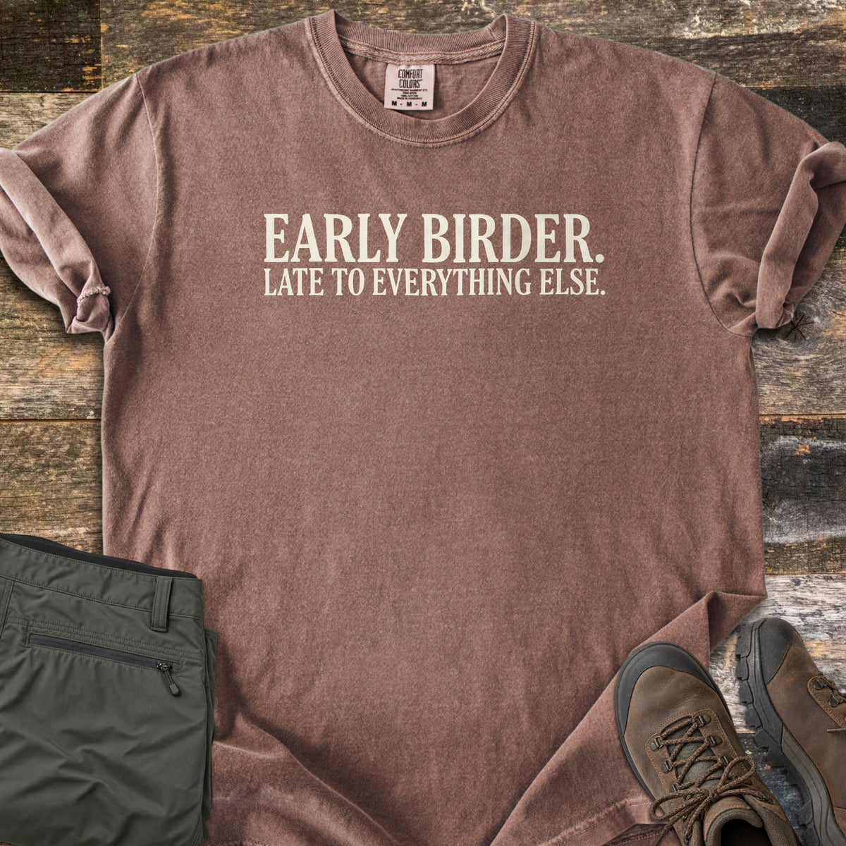 Early Birder Comfort Colors T-shirt in Espresso, laid flat. Cream serif text across the chest reads EARLY BIRDER. LATE TO EVERYTHING ELSE. The shirt has rolled sleeves and lies on weathered wood planks beside folded olive hiking pants and brown hiking boots.