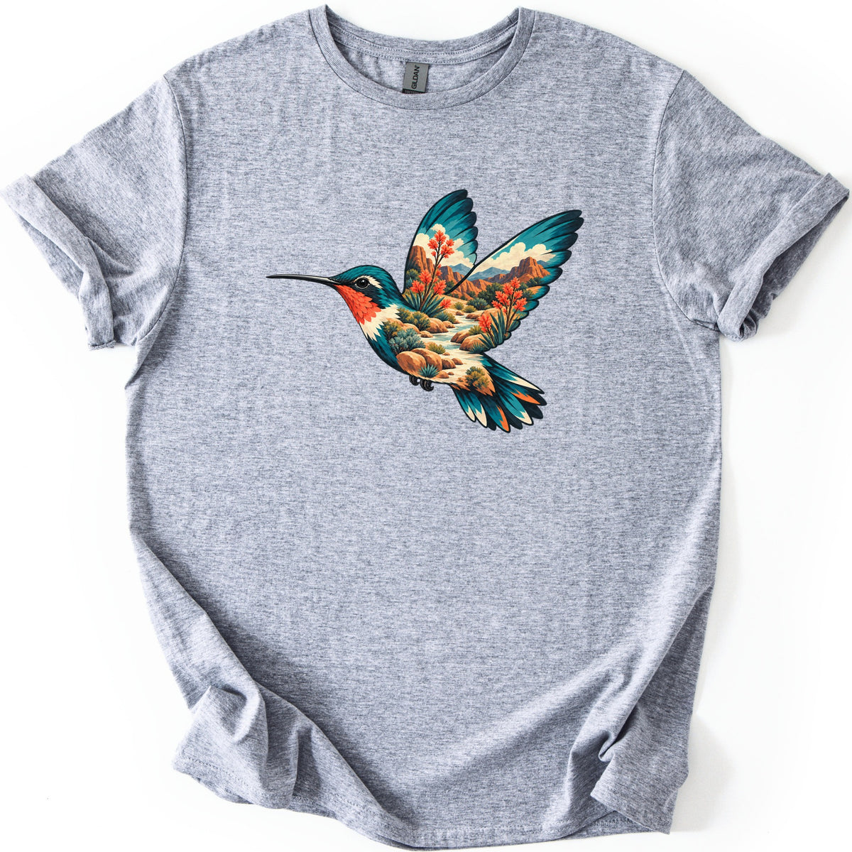 Hummingbird Canyon Comfort Colors T-shirt in Sport Grey, laid flat. A teal and red-throated hummingbird in flight, its body and wings showing a desert canyon with red cliffs, white clouds, a winding creek, boulders and orange-red flowers. Sleeves cuffed, on white.