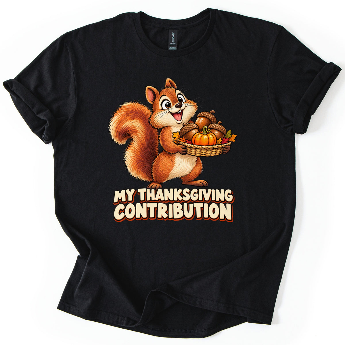 My Thanksgiving Contribution Comfort Colors T-shirt in Black, laid flat. A grinning cartoon squirrel with a bushy tail holds a basket of acorns, a pumpkin and fall leaves above the words My Thanksgiving Contribution in cream letters. The shirt lies on a white surface with cuffed sleeves.