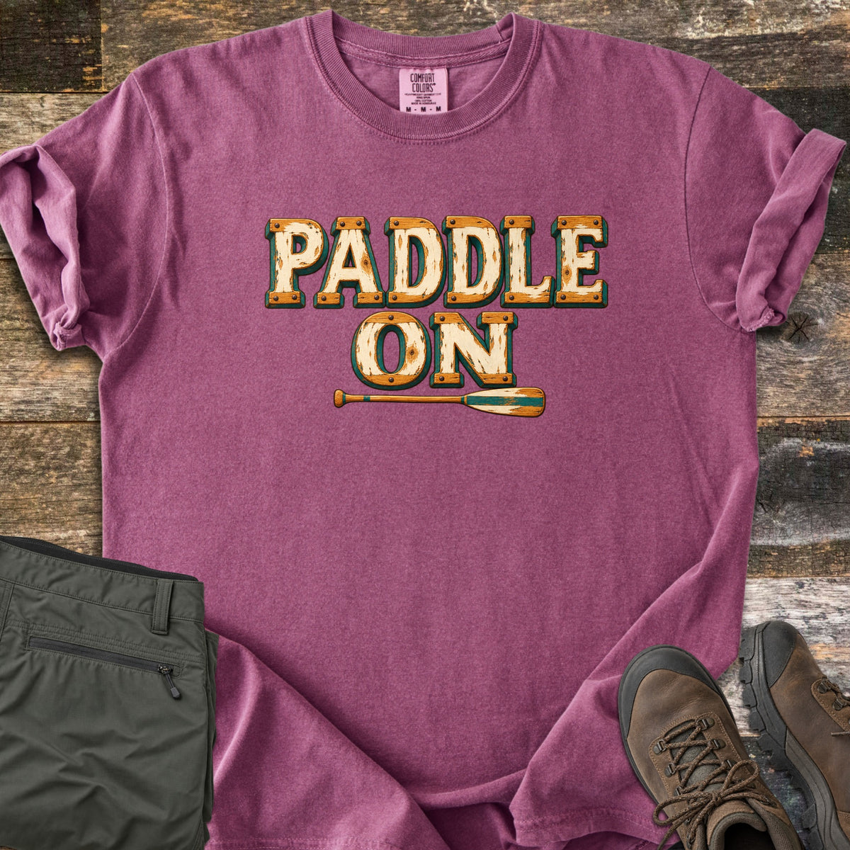 Paddle On Comfort Colors T-shirt in Berry, laid flat. The words Paddle On in distressed cream lettering with wood-grain edges and rivets, above a wooden canoe paddle. Folded green hiking pants and brown hiking boots sit on a weathered wood floor.