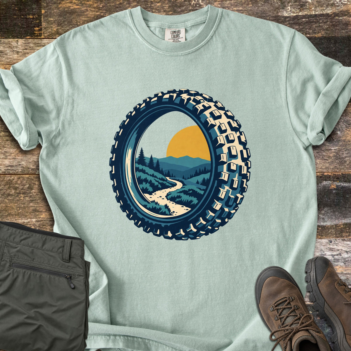 Knobby Tire Trail Comfort Colors T-shirt in Bay, laid flat. A mountain bike tire frames a scene of a winding trail through green hills and pine trees toward mountains and a yellow sun. Olive cargo pants and brown hiking boots lie beside it on weathered wood planks.