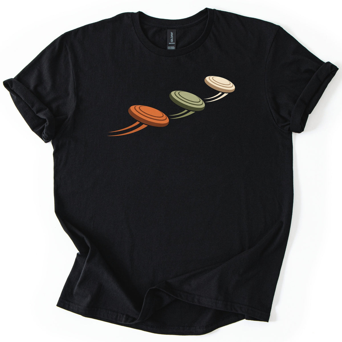 Flying Clays Comfort Colors T-shirt in Black, laid flat. Three clay targets in orange, olive green and cream fly upward in a diagonal line across the chest, each trailing motion streaks, with no words. The shirt lies on a white surface with cuffed sleeves.