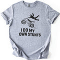 ATV Stunts Comfort Colors T-shirt in Sport Grey, laid flat. A black silhouette of a helmeted rider flying off the back of an ATV as it kicks up dirt, above the words I DO MY OWN STUNTS. The shirt lies on a white background with its sleeves cuffed.