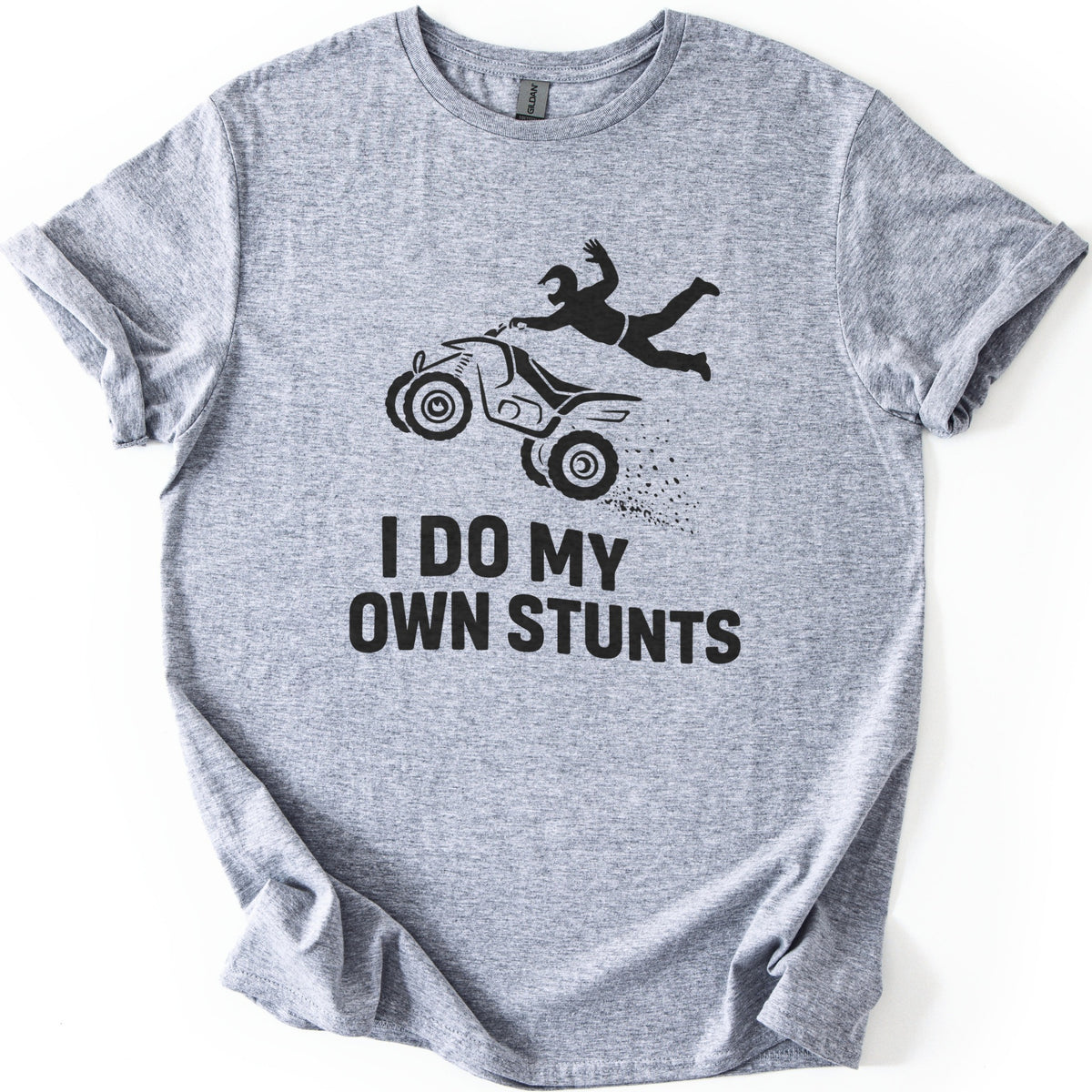 ATV Stunts Comfort Colors T-shirt in Sport Grey, laid flat. A black silhouette of a helmeted rider flying off the back of an ATV as it kicks up dirt, above the words I DO MY OWN STUNTS. The shirt lies on a white background with its sleeves cuffed.