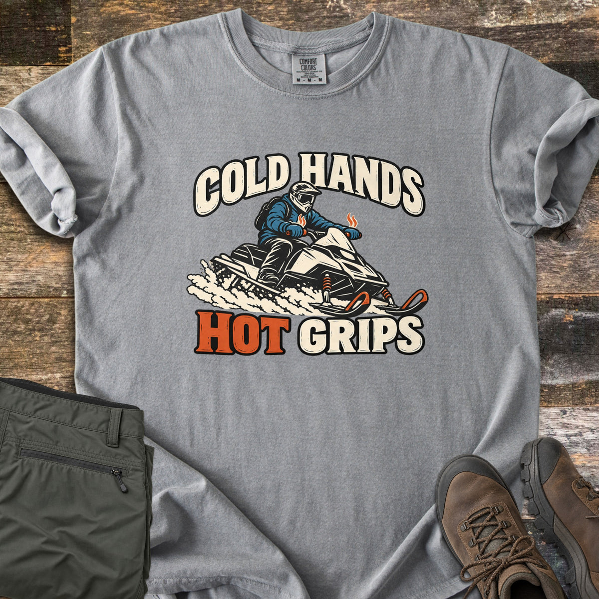 Cold Hands Hot Grips Comfort Colors T-shirt in Grey, laid flat. A snowmobile rider in a helmet and goggles kicks up snow, with steam rising from the handlebar grips, between the words Cold Hands and Hot Grips. Shown on rustic wood with olive cargo pants and brown hiking boots.