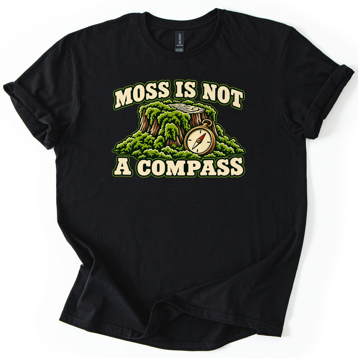 Moss Is Not a Compass Comfort Colors T-shirt in Black, laid flat. Cream lettering reads Moss Is Not above a tree stump covered in green moss, with a brass pocket compass beside it, and A Compass below. The shirt lies on a white background, sleeves cuffed.
