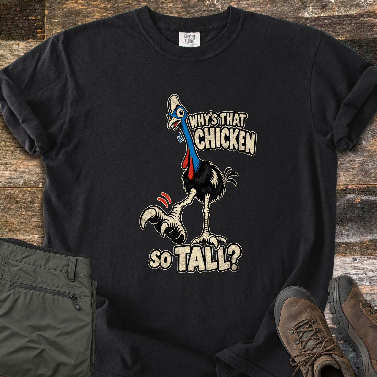 Why's That Chicken So Tall Comfort Colors T-shirt in Black, laid flat. A cartoon cassowary with a blue neck, red wattles and a huge clawed foot raised mid-step, with the words Why's That Chicken So Tall? Folded green cargo pants and brown hiking boots sit on weathered wood.