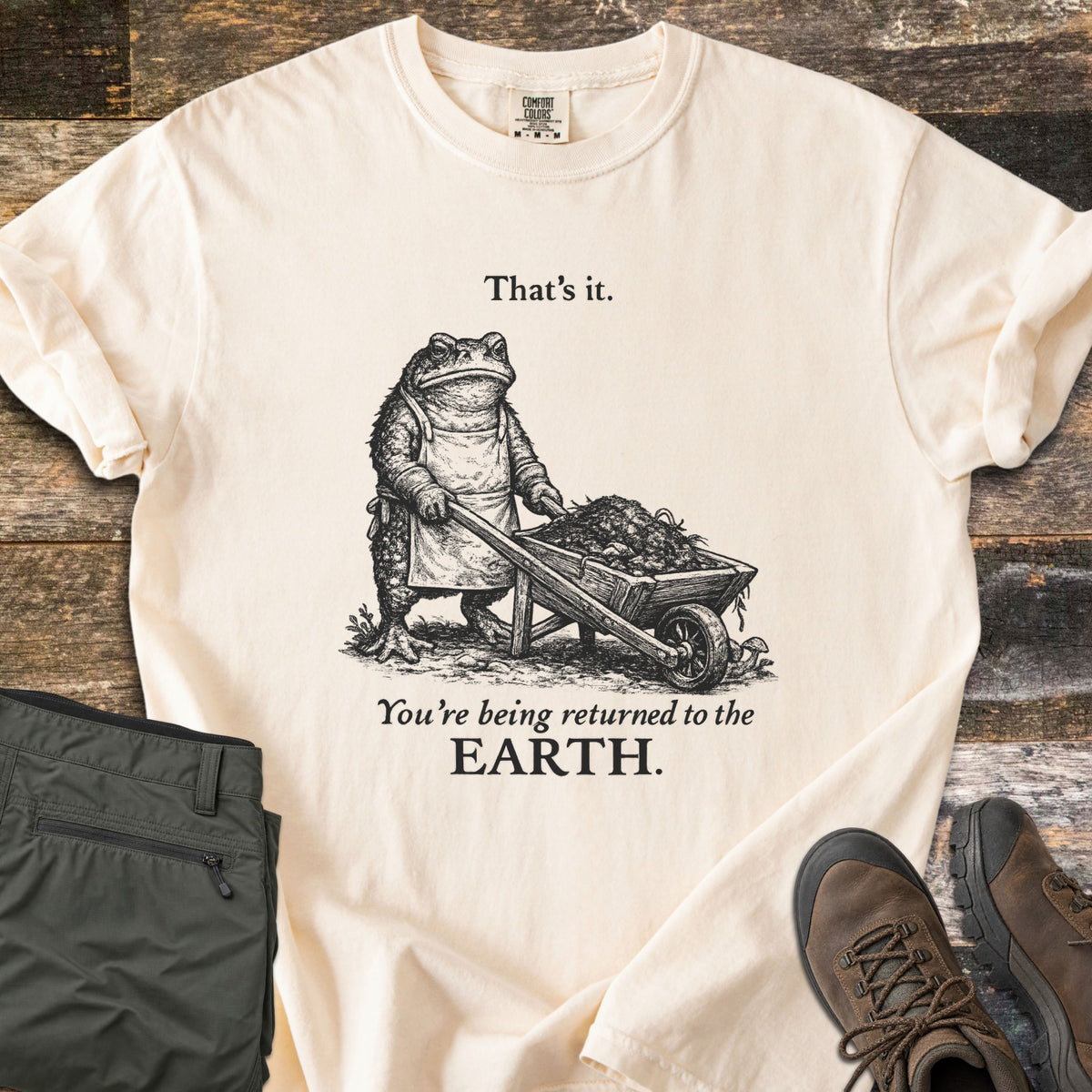 Toad Compost Comfort Colors T-shirt in Ivory, laid flat. A black-ink toad in an apron pushes a wheelbarrow of compost between the words That's it. and You're being returned to the EARTH. Folded olive pants and brown hiking boots sit beside it on rustic wood planks.