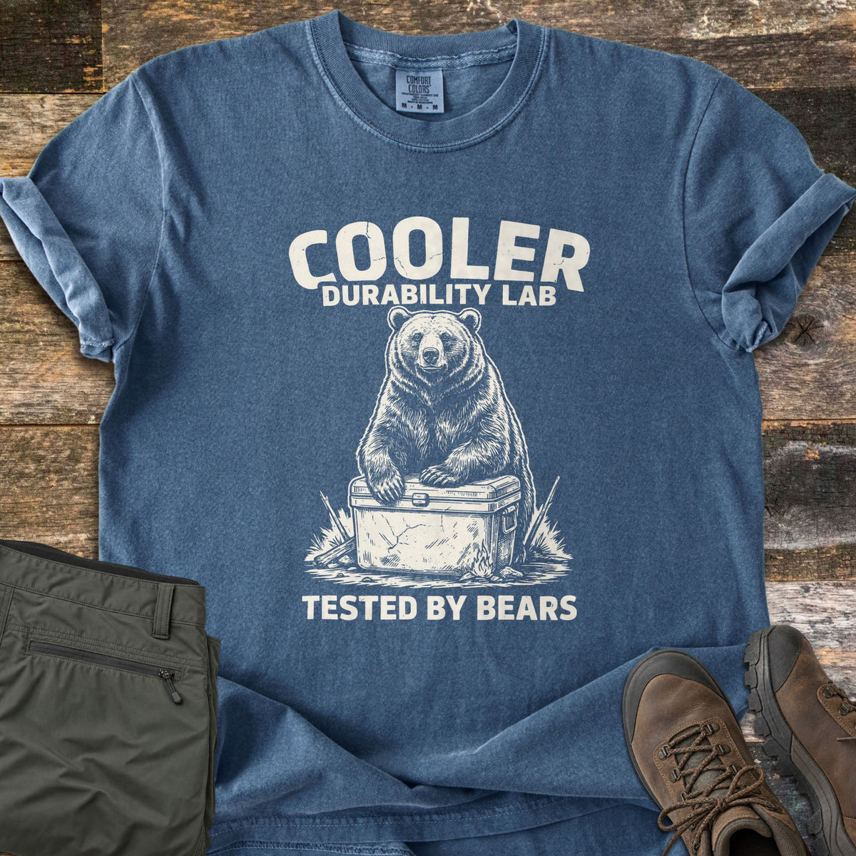 Cooler Durability Lab Comfort Colors T-shirt in Denim, laid flat. A sketched bear rests its paws on a cracked cooler above the words Cooler Durability Lab and Tested By Bears. Sleeves are rolled on a wood plank floor beside olive cargo pants and brown hiking boots.