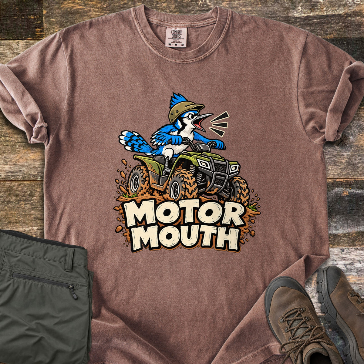 Motor Mouth Comfort Colors T-shirt in Espresso, laid flat. A cartoon blue jay in a tan helmet, beak wide open, rides a green ATV kicking up mud above the words Motor Mouth. Folded olive pants and brown hiking boots rest beside it on rustic wood boards.