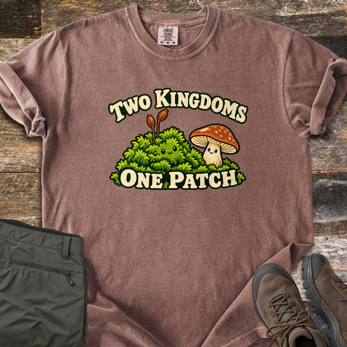 Two Kingdoms One Patch Comfort Colors T-shirt in Espresso, laid flat. A smiling moss clump with two orange stalks sits beside a smiling spotted brown mushroom, between the words Two Kingdoms and One Patch. Sleeves rolled, on weathered wood with olive hiking pants and brown boots.