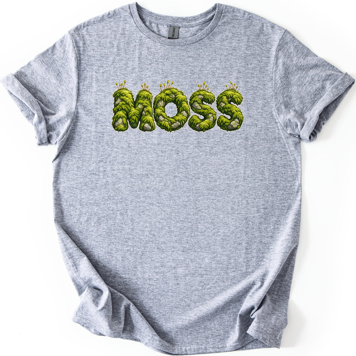 Moss Comfort Colors T-shirt in Sport Grey, laid flat. Across the chest, the word MOSS is spelled in rounded letters covered in green moss, with gray stones and small sprouts on top. The shirt lies on a white background with sleeves cuffed.