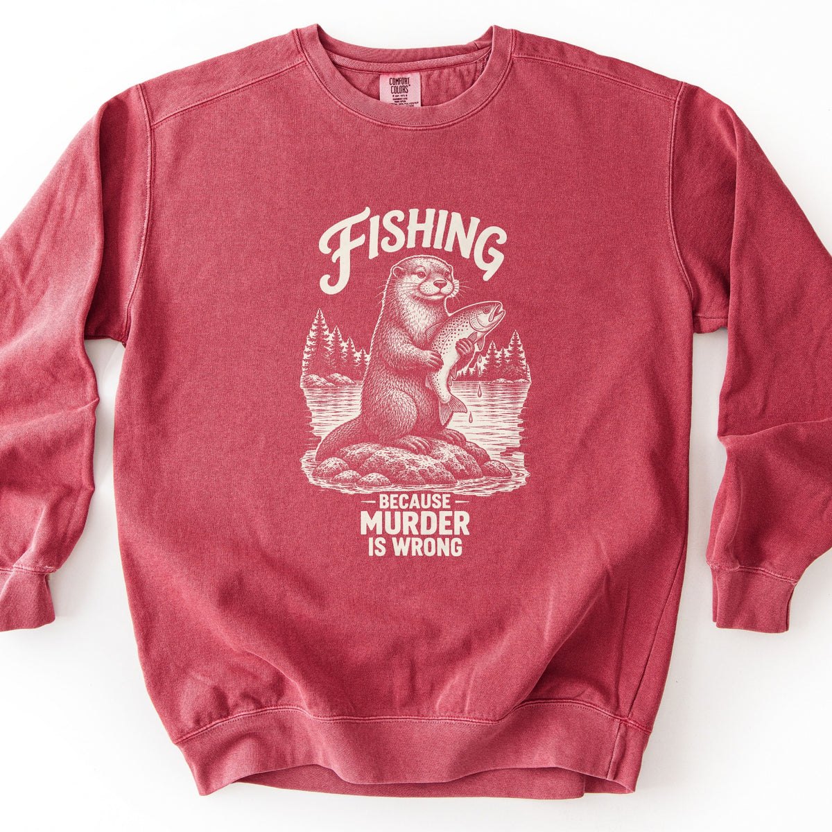 Fishing Because Murder Is Wrong Comfort Colors crewneck sweatshirt in Crimson, laid flat. A cream sketch-style otter sits on a rock in a lake holding a dripping trout, with pine trees behind it. Fishing arches above it and Because Murder Is Wrong is below. Set on a white surface.
