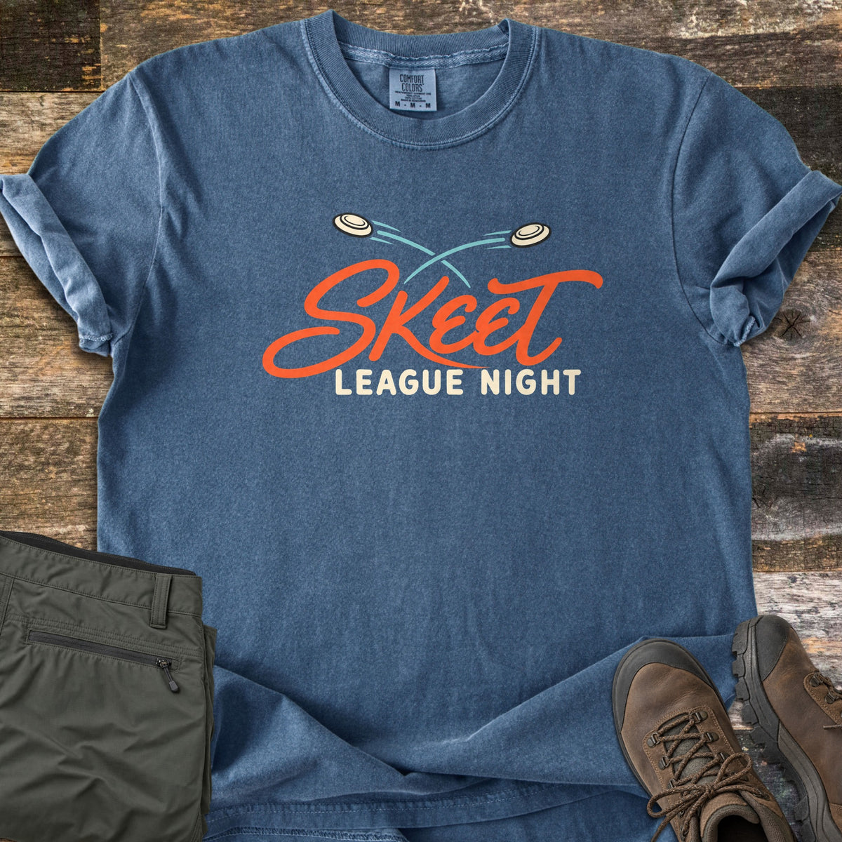 Skeet League Night Comfort Colors T-shirt in Denim, laid flat. Two clay targets fly across teal trails above the words Skeet League Night in orange script and cream letters. It lies on weathered wood with olive cargo pants and brown hiking boots.