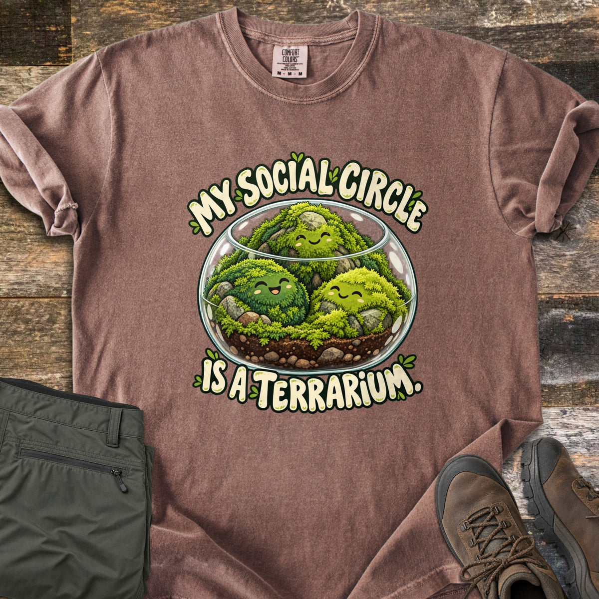 My Social Circle Is a Terrarium Comfort Colors T-shirt in Espresso, laid flat. A glass bowl terrarium holds three smiling moss balls among rocks and soil, between the words My Social Circle and Is a Terrarium. Folded olive pants and brown hiking boots sit on weathered wood planks.