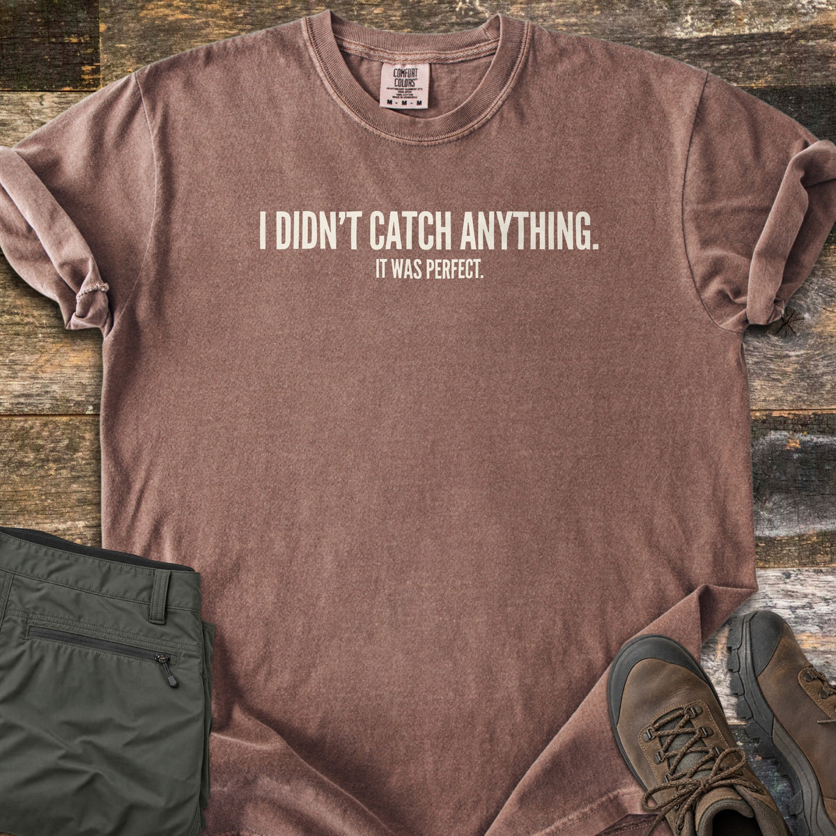 Didn't Catch Anything Comfort Colors T-shirt in Espresso, laid flat. Off-white block letters read I Didn't Catch Anything. with It Was Perfect. in smaller letters below. Sleeves are rolled, with folded olive hiking pants and brown hiking boots on weathered wood planks.