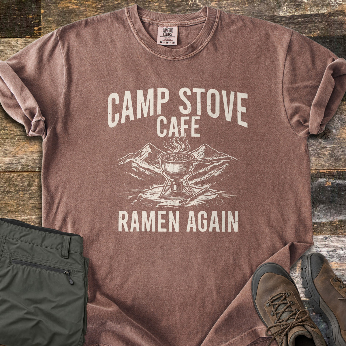 Camp Stove Cafe Comfort Colors T-shirt in Espresso, laid flat. Printed in cream: a steaming pot on a camp stove atop a rock with mountains behind, under the words Camp Stove Cafe and above Ramen Again. Sleeves rolled, on weathered wood with green pants and hiking boots.